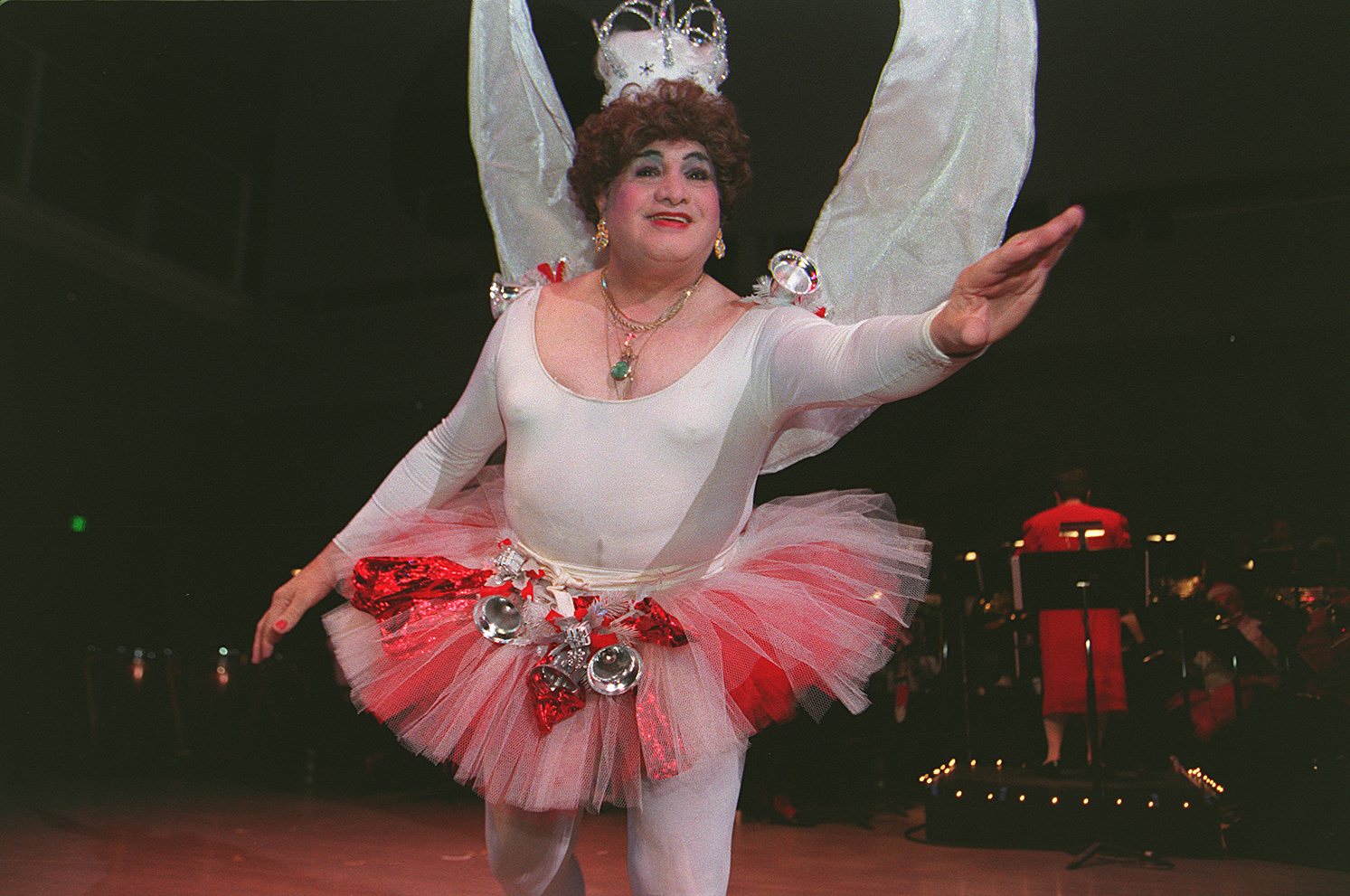 A person wearing a white full body leotard and a pink tutu and white angel wings and a crown. They are gesturing toward the camera, as if to take flight.