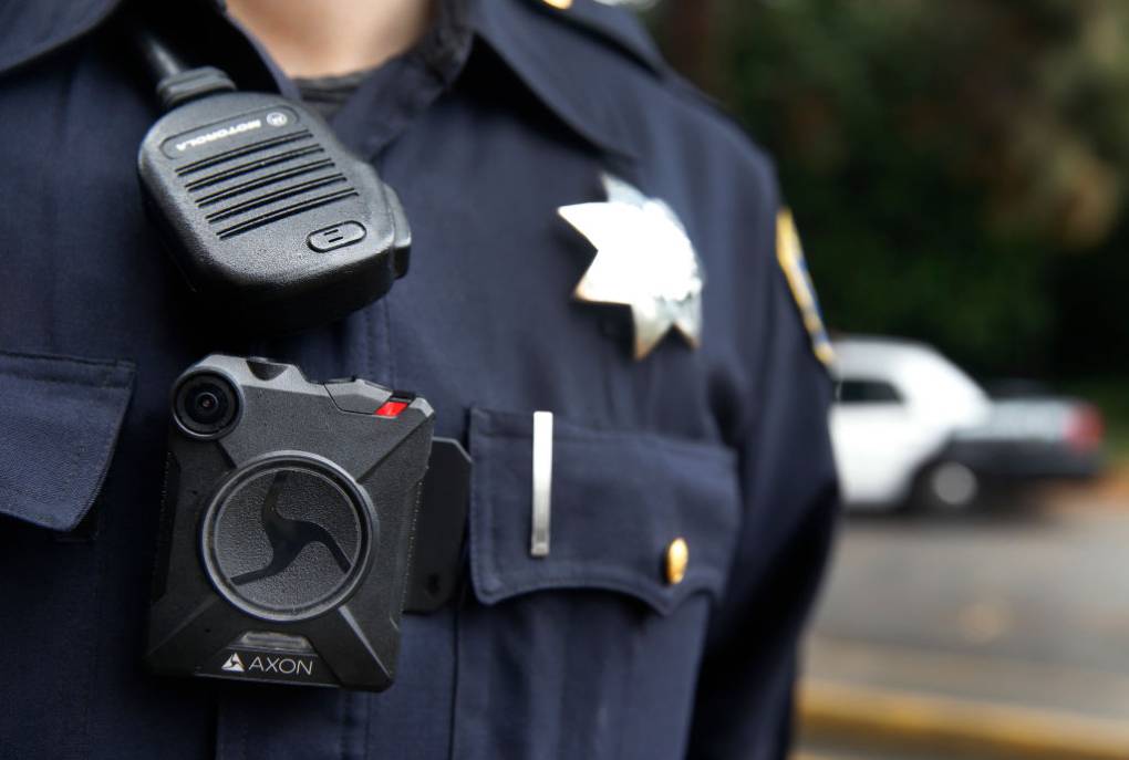 The torso of a police officer wearing a bodycam, a walkie-talkie, a badge, and a pen in a pocket, with a blurred white car in the background.