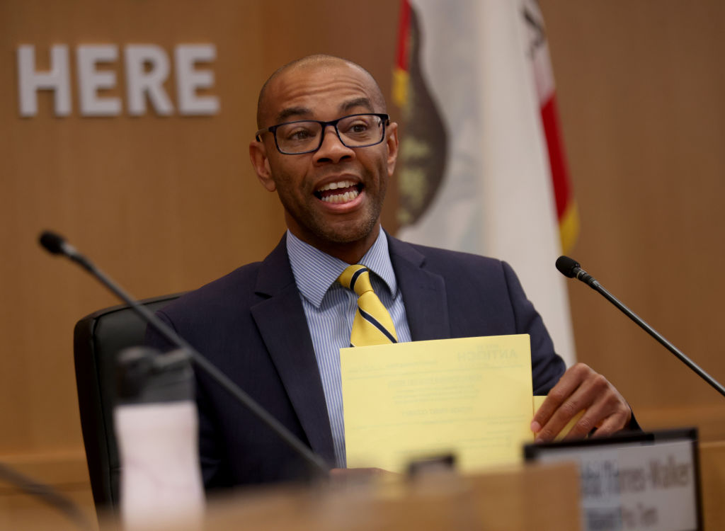 An African American man wearing a suit and tie speaks in a city council meeting.