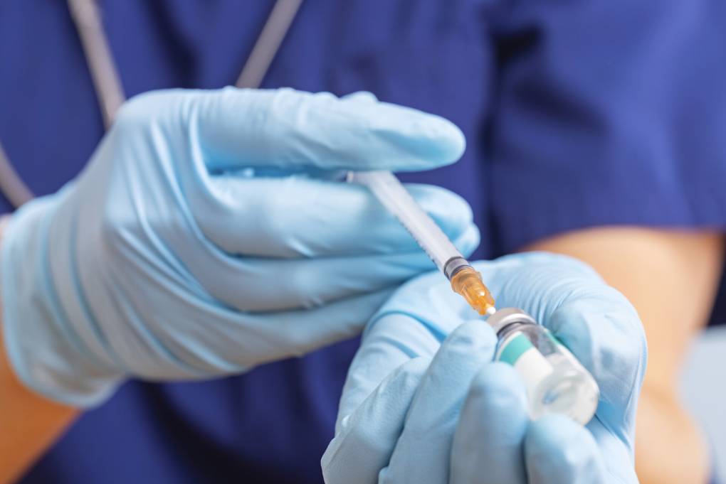 A pair of gloved hands fills a syringe from a vial of vaccine.