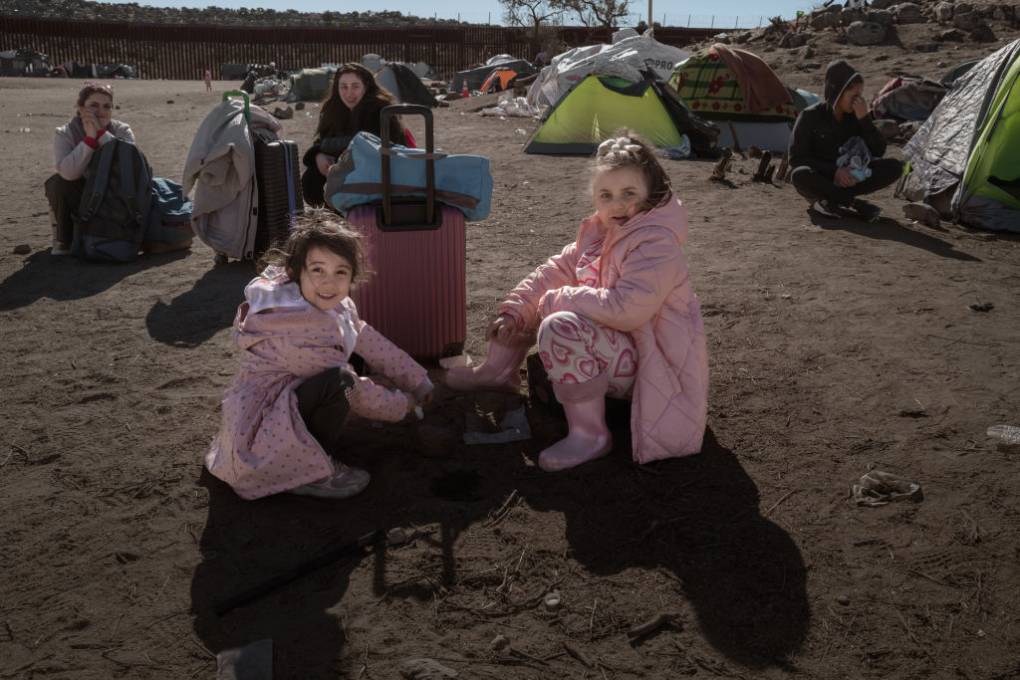 Two small girls in pink coats sit outside a tent with people in the background.