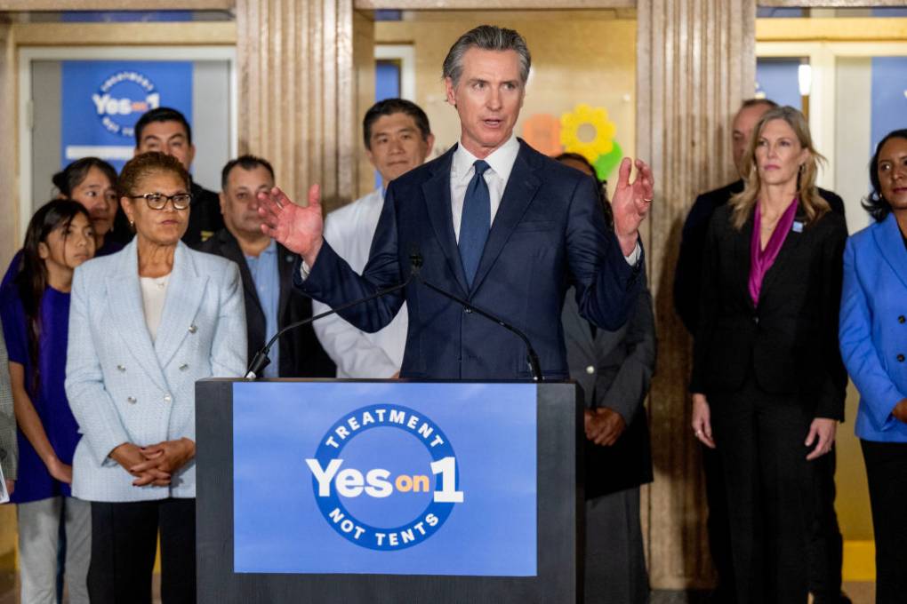 A white man in a blue suit and blue tie raises his hands in gesture as he speaks from behind a lectern with "Treatment, Not Tents, Yes on 1" written on it and people standing behind him listening.