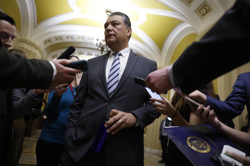 Sen. Alex Padilla stands center frame surrounded by reporters pointing microphones at him.