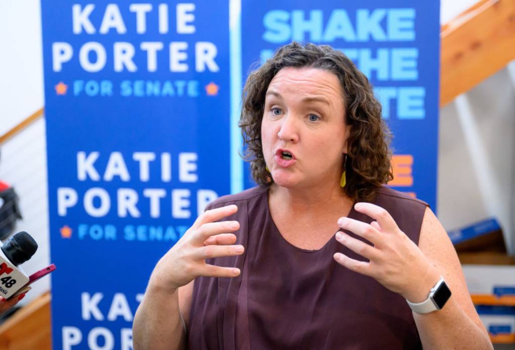A woman wearing a sleeveless red shirt has her hands raised with signs that read "Katie Porter for Senate" in the background.