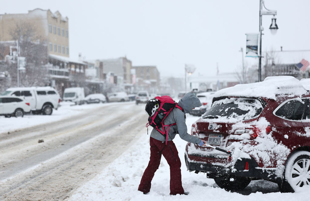 A person behind their SUV on the side of a snow-covered road ion a snow covered town and street.