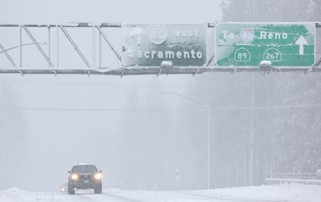 A car with headlights on through a snowstorm and roads covered in snow, with a traffic sign above that reads Sacramento and Reno.