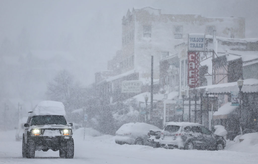 Snowfall covering a street, buildings and cars.