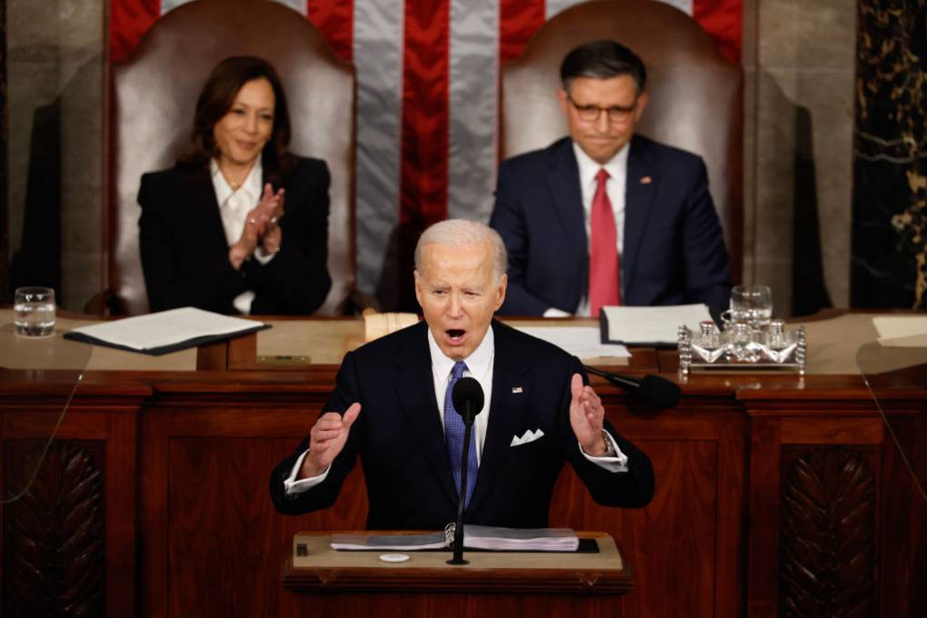President Joe Biden delivers a speech, with hands raised, as Vice President Kamala Harris and another man stand behind him.