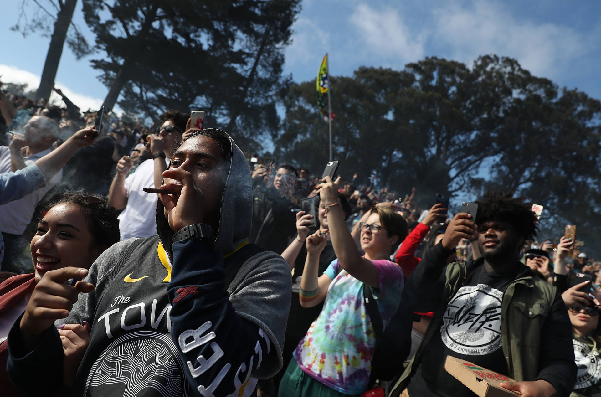 A crowd of festivalgoers with some dancing and smoking outdoors, with trees in the background.
