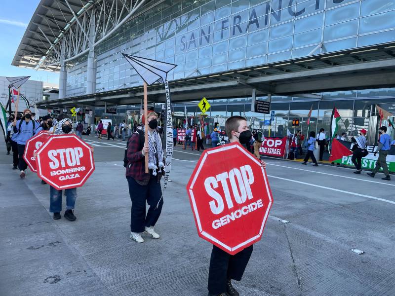 Protesters stand outside SFO holding signs that say 'Stop Genocide.'