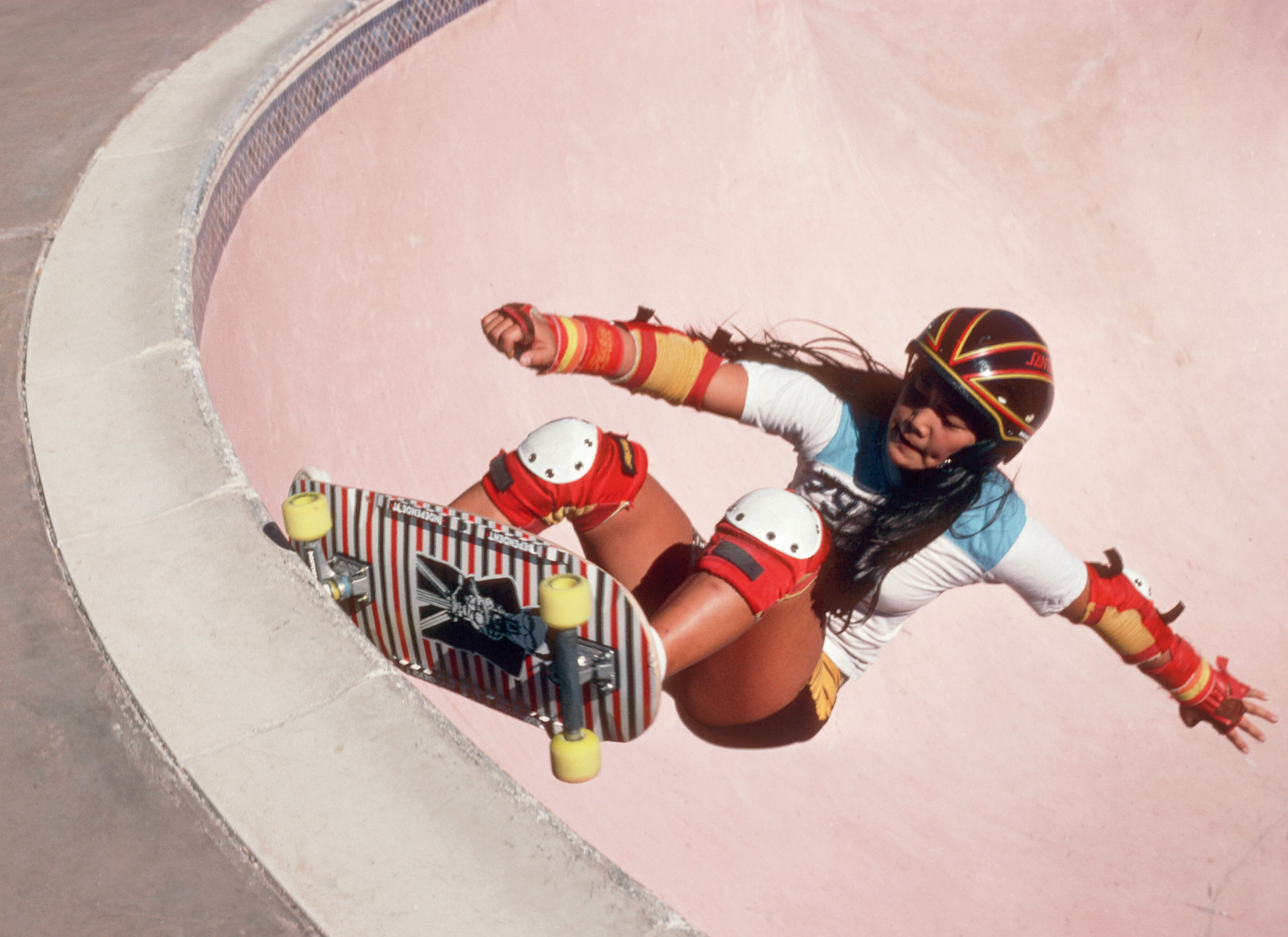 A young woman skateboarder with helmet and knee pads skates a ramp in an old photo.
