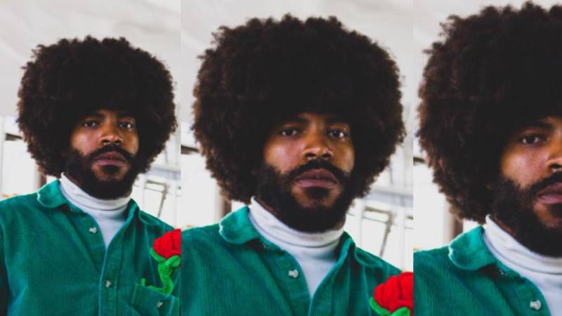 Portrait of a young Black man with a afro and beard in a white turtleneck shirt with a green button up shirt on top of it.