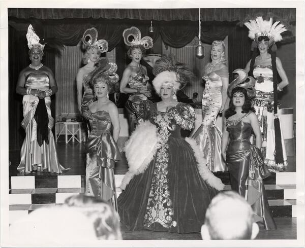 A black and white photo featuring eight drag queens posing on a multi-tiered stage, wearing gowns.