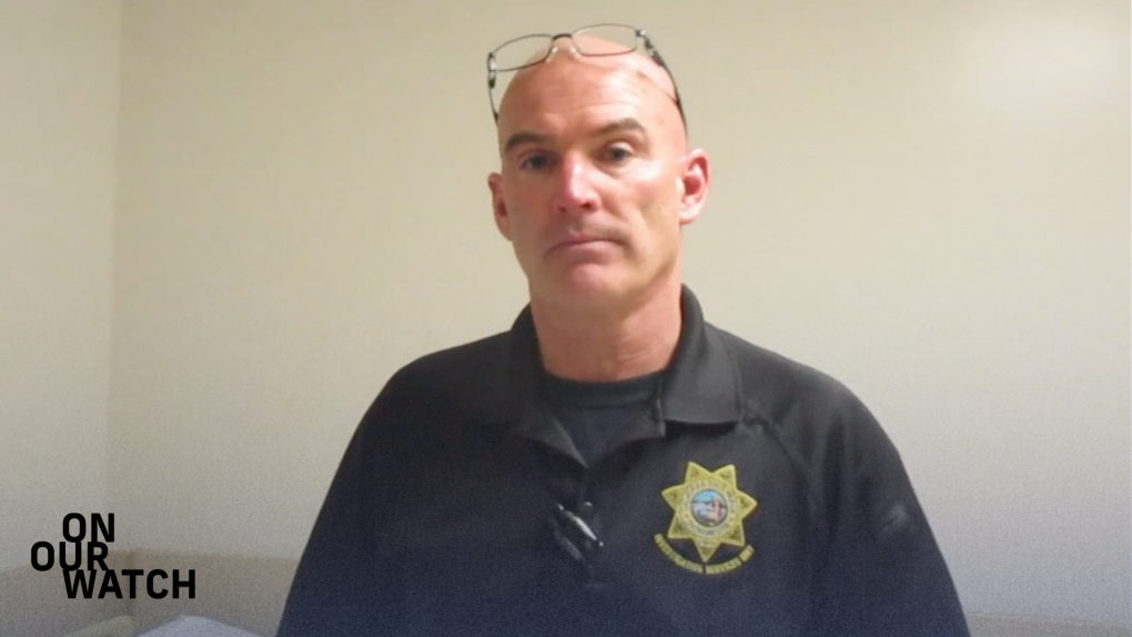 A white man with a shaved head and wearing a black uniform shirt with a gold "Investigative Services Unit" insignia stares at the camera. His glasses are perched on top of his head and he is standing in a nondescript beige room.