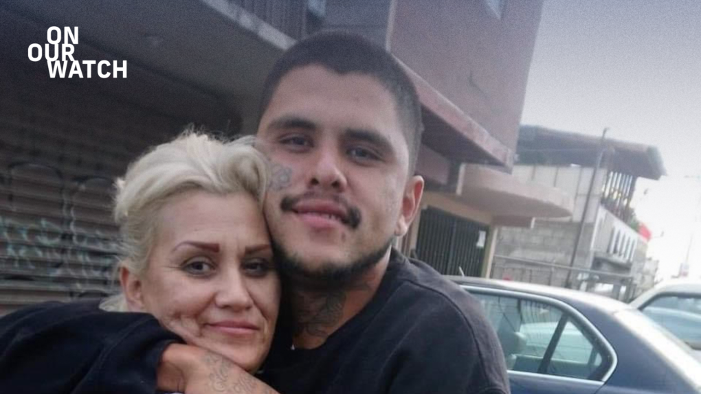 A young Latino man with short black hair, a mustache and tattoos on his neck and face looks at the camera. He has his arm around his mother, a youthful-looking middle-aged Latina woman with platinum blonde hair. His cheek is pressed to the side of her head and they’re both smiling with mouths closed. They are standing in front of several parked cars and a brown building with graffiti on a metal roll-up door.