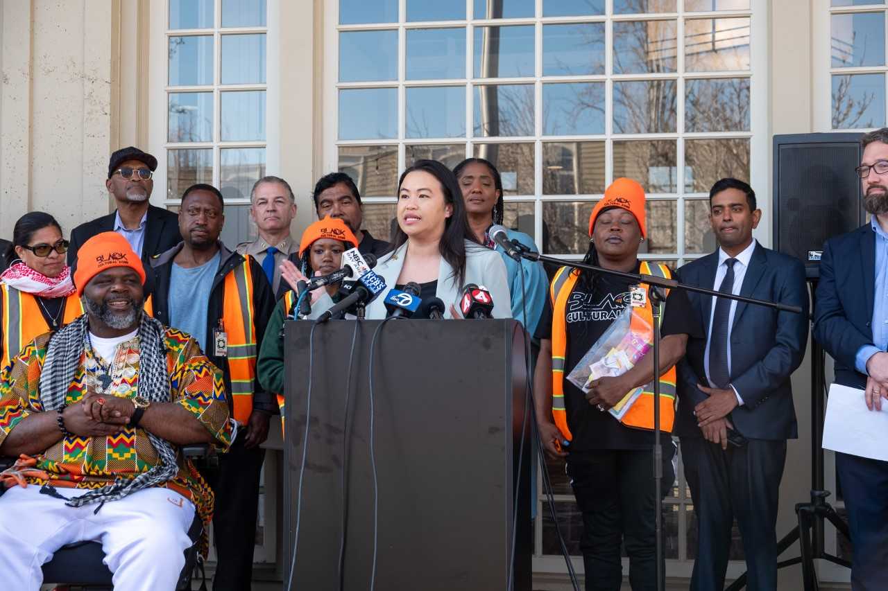 An Asian woman stands at a podium surrounded by people in safety vests and suits.