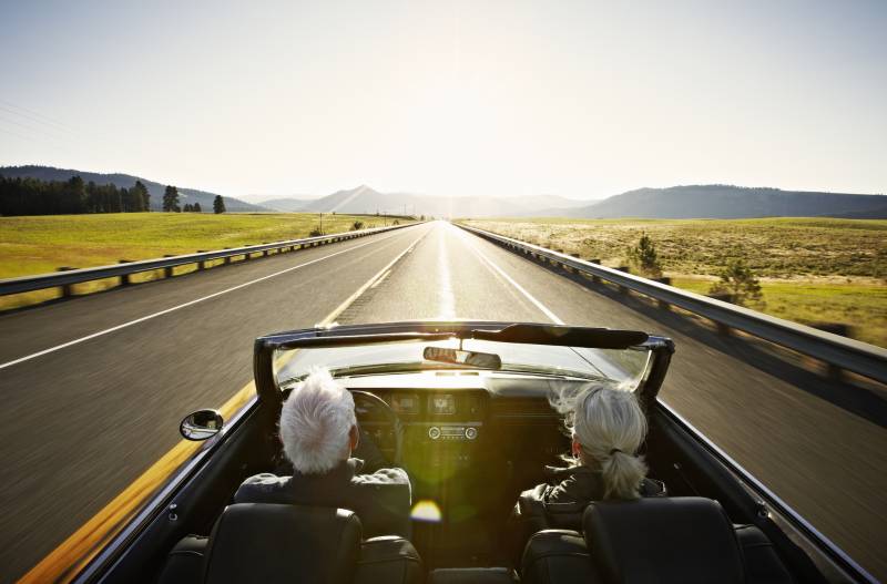 A senior couple driving convertible car at sunrise on rural highway.