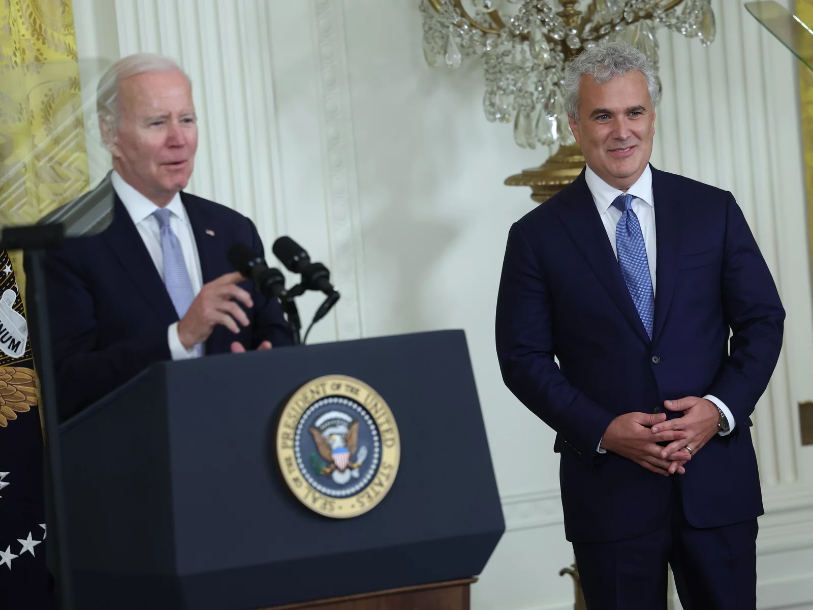 Two white men in blue suits with blue ties, one standing behind a lectern with the emblem of the President ofd the UNited States on it.