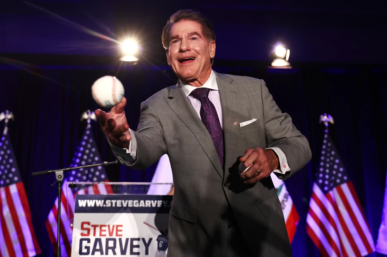 A white man in a grey suit with a purple tie throws a baseball into the crowd with American flags on the stage behind him.