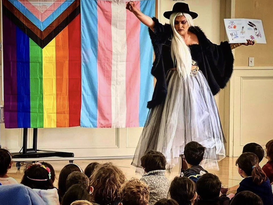 A drag queen stands, gesturing dramatically while reading from a book. A handful of children sit by her feet.