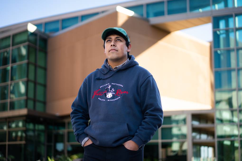 A man stands outside a building while wearing a hat and dark blue sweatshirt with his hands in his pockets.