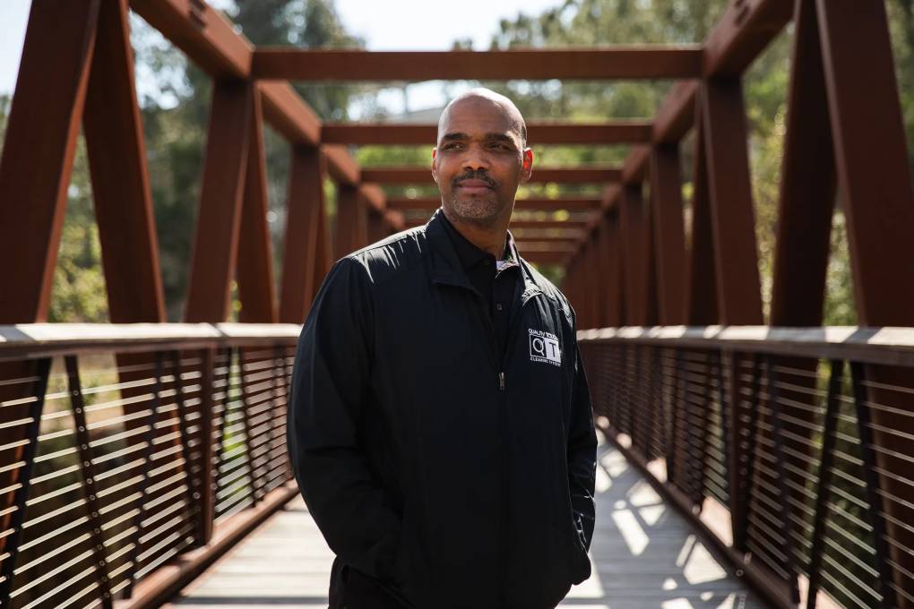 A Black man in a cleaning uniform stands on a bridge.