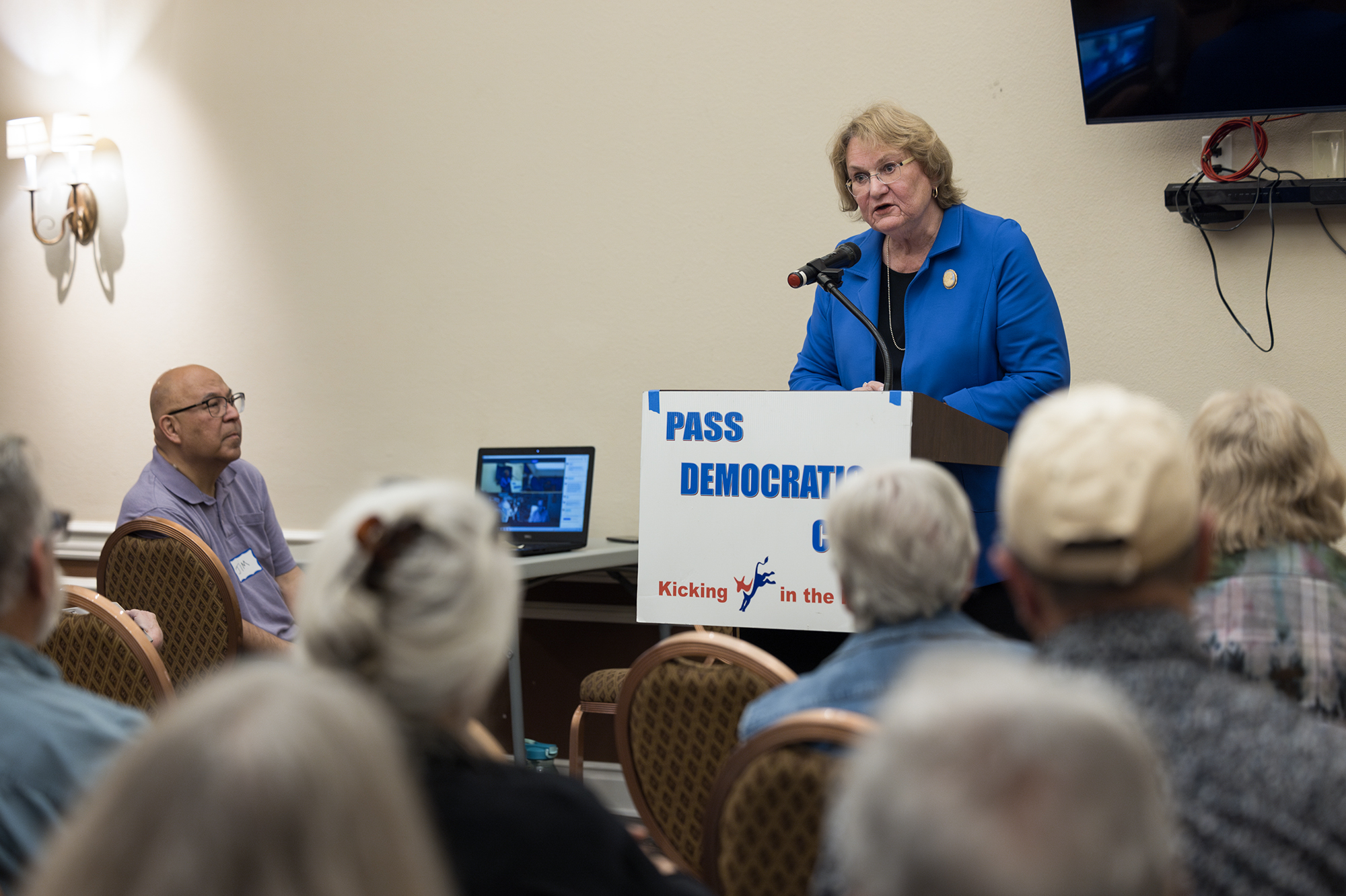 A white woman wearing glasses and a blue jacket stands behind a microphone and podium in front a group of people who are seated.