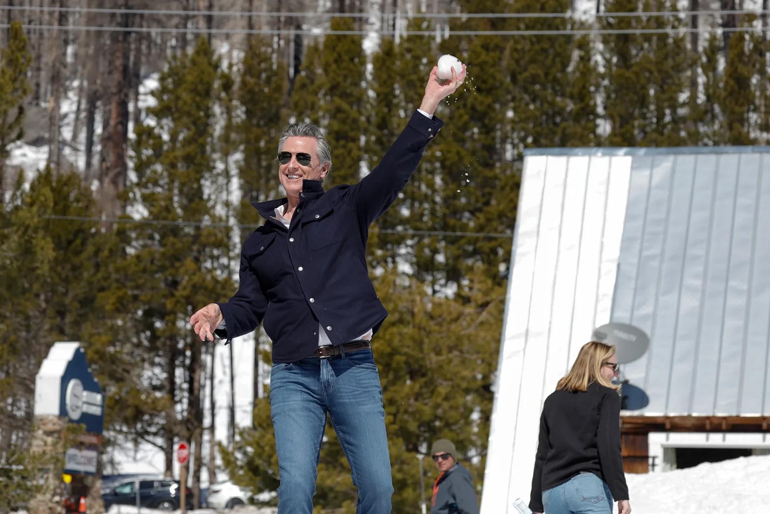 A middle-aged white man wearing jeans and a blue buttoned shirt and sunglasses throws a snowball with snow and trees in the background.