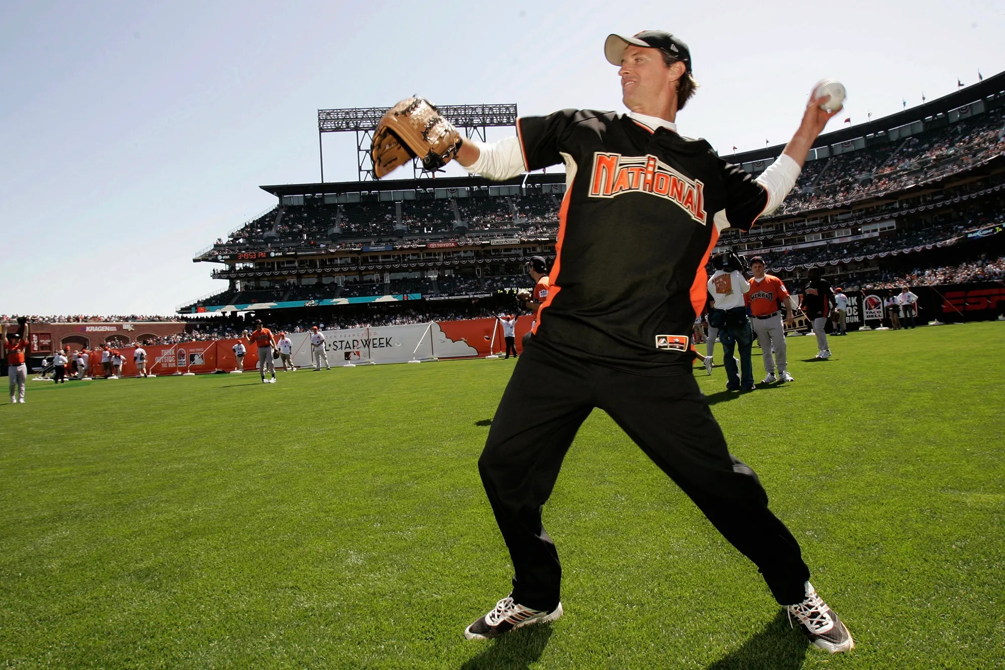 A middle aged man pitches a baseball in a full ballpark and wearting a "nationals" outfit.