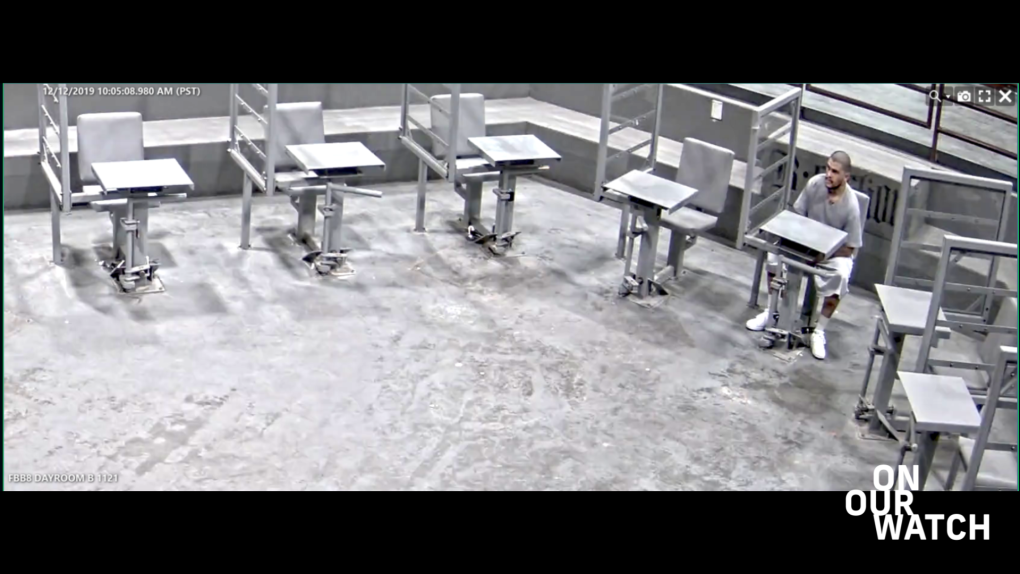 A screenshot of prison surveillance footage shows a gray room with a cement floor. On the right side of the image sits a Latino man in a gray shirt and white shorts with a goatee and a buzz cut, or shaved head. His legs are chained to the metal desk he’s sitting at, and he’s staring across the room towards the left of the image. Positioned around him at regular intervals are six more chair-desk combos as well as metal dividers between them.