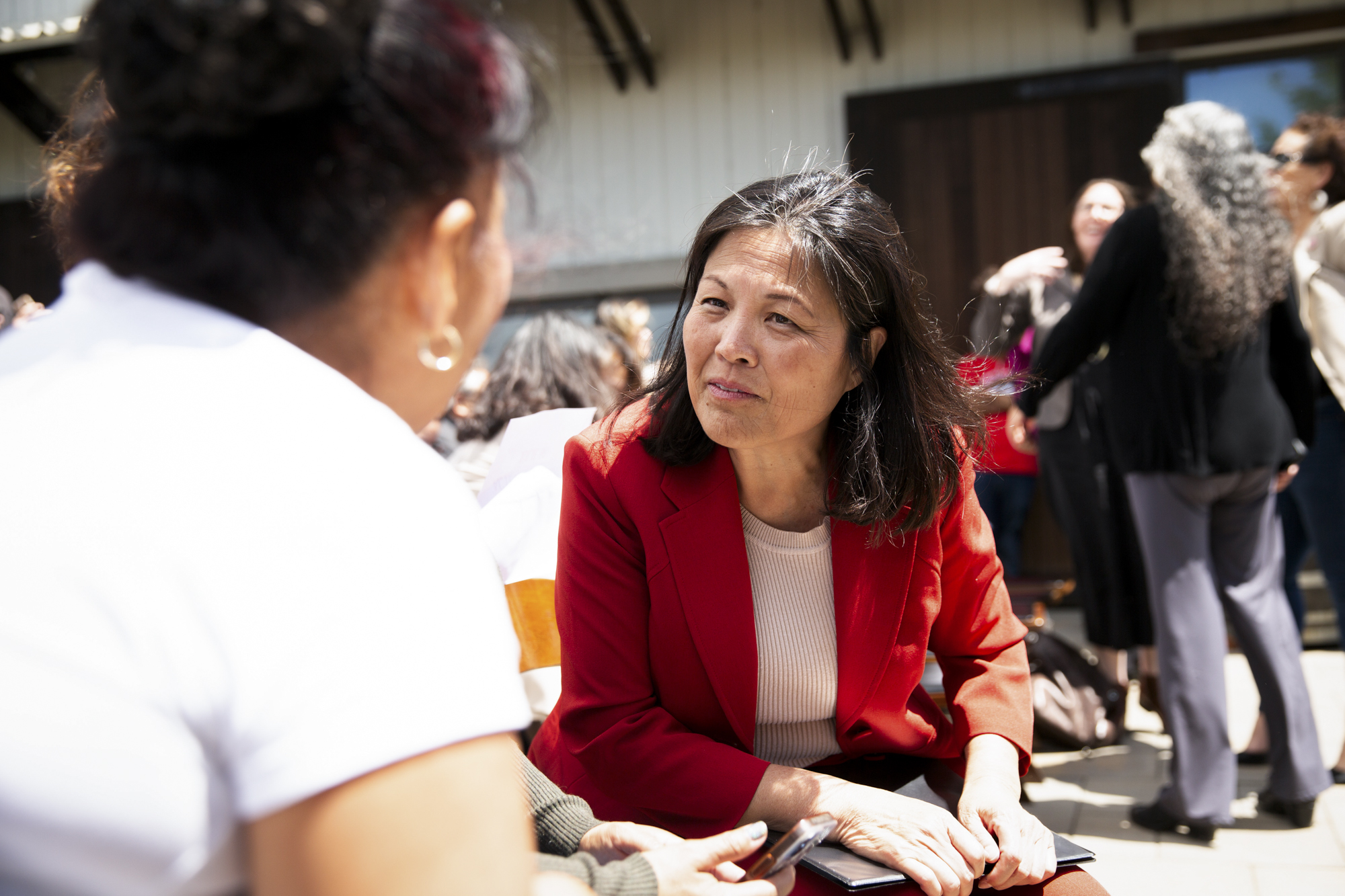 An older Asian woman in a red suit leans over as she speaks to a woman seen from behind.