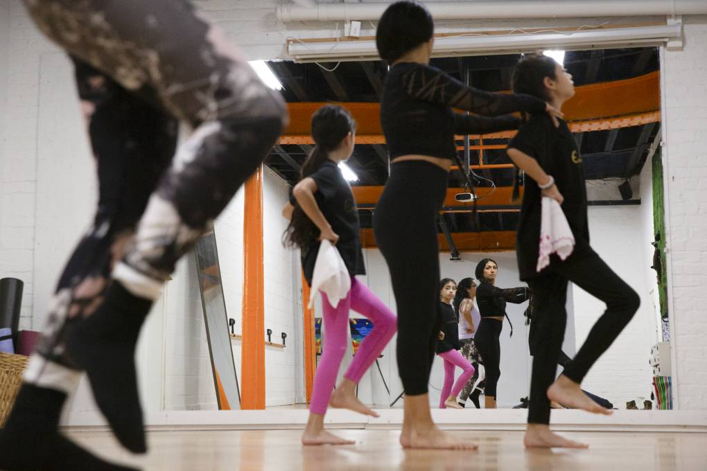 Several women of all ages practice dance moves in a mirror at a dance studio.