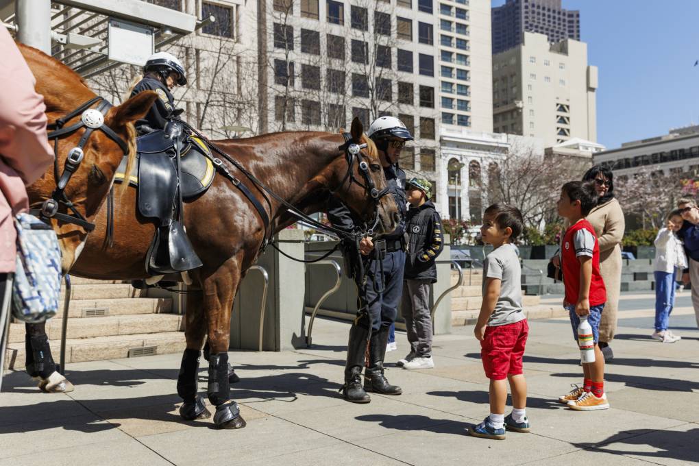 Kids interact with police officers on horseback in an urban plaza.