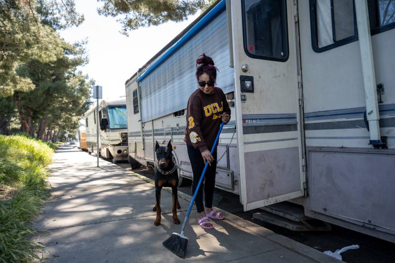 A woman sweeps the sidewalk near an RV while a dog stands nearby.