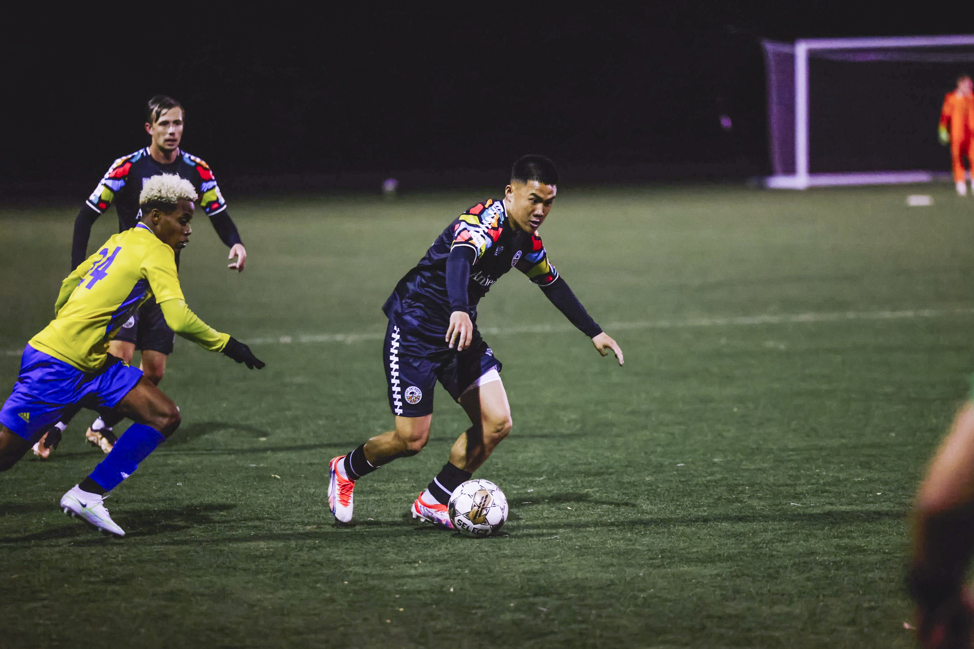 Three men wearing soccer equipment and kits play on a field with a goalie in the background.