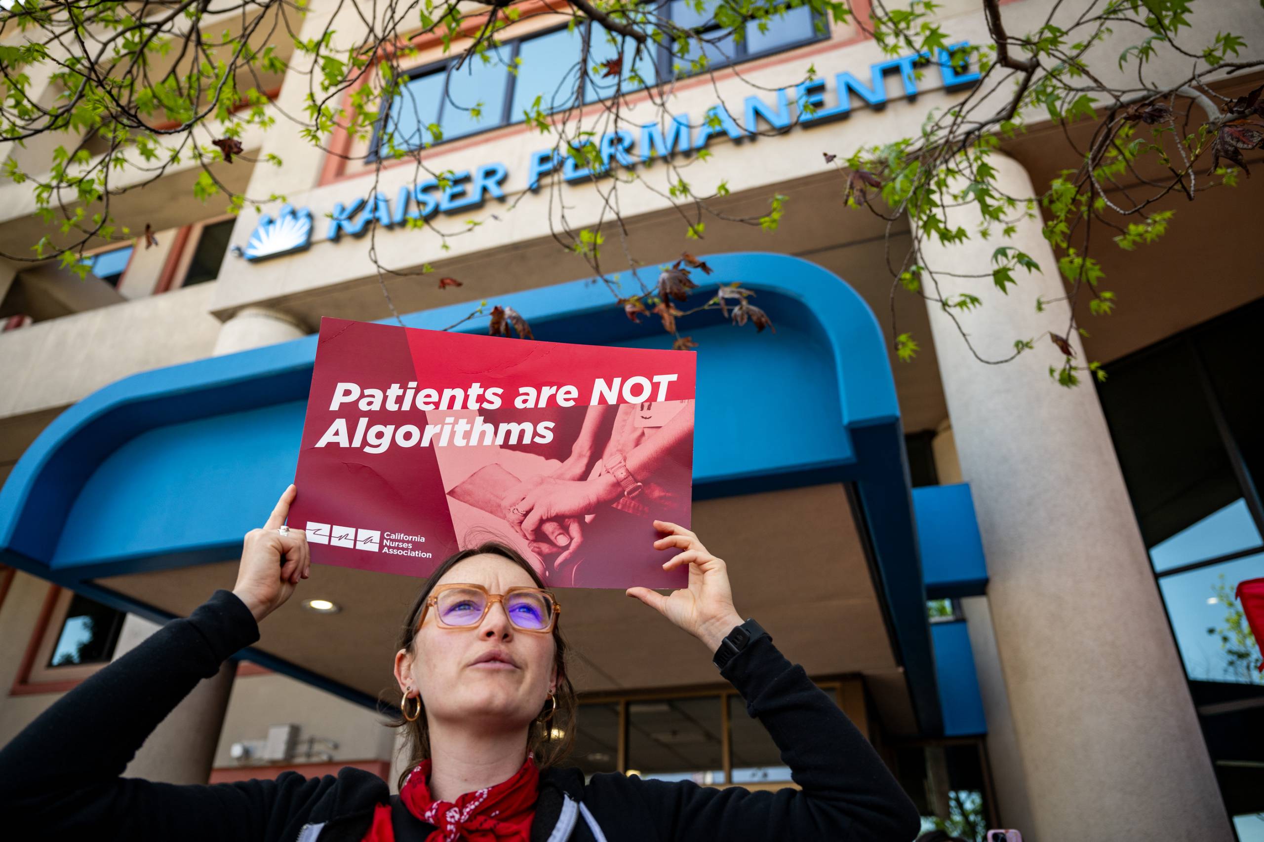 A woman with glasses holds a sign above her head that reads 'Patients are NOT algorithms' in front of a Kaiser Permanente building.