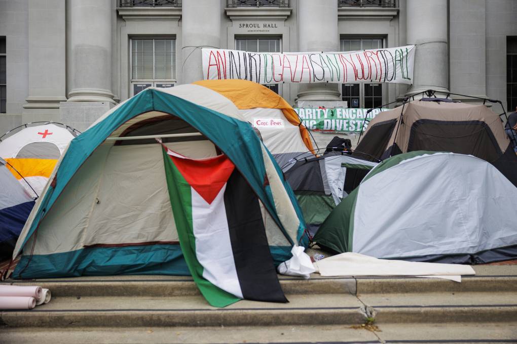 Tents set up on the steps leading up to a building. A red-white-green-black Palestinian flag hangs from one tent. One banner hung from the building in the background says: 'An injury to Gaza is an injury to us all.' Another says: 'Divest.'