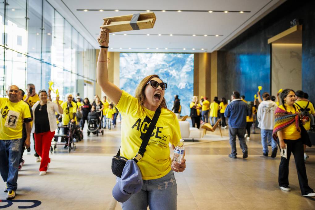 A woman wearing sunglasses and a yellow shirt holds up an object in one hand and a water bottle in the other with people in the background.