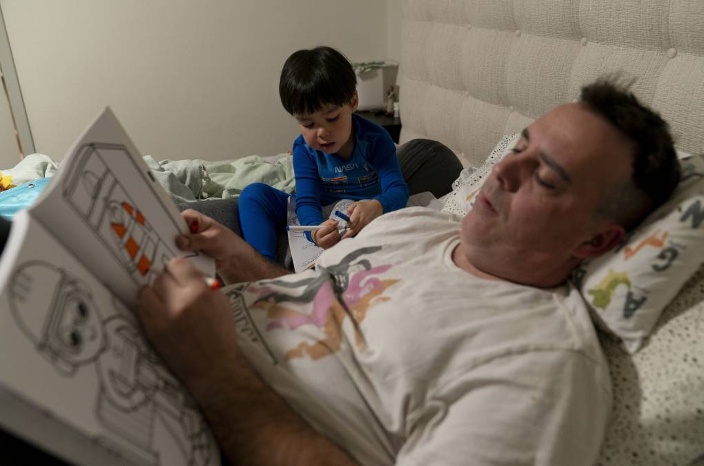 A man lies in bed reading a book while his young son sits next to him listening.