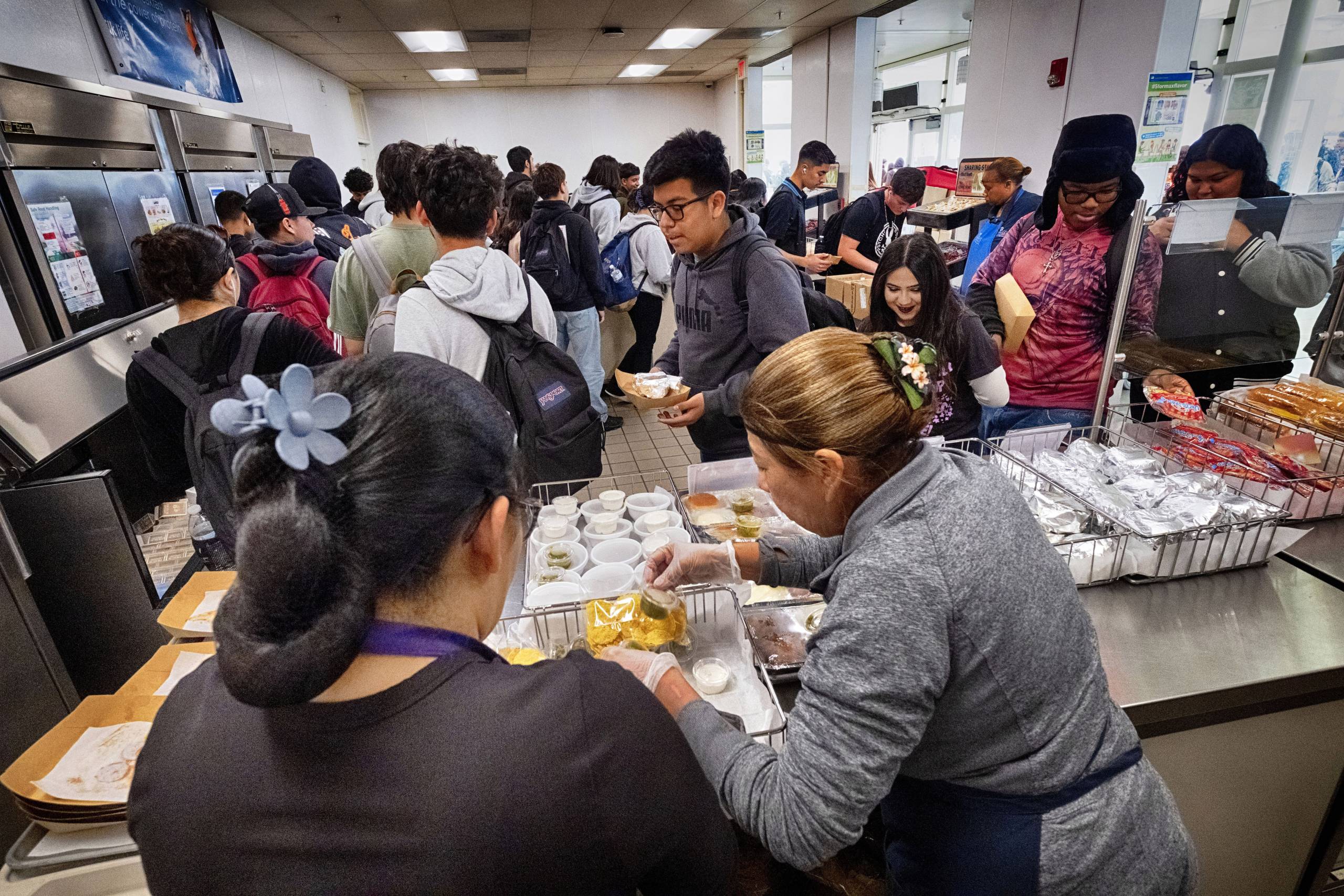 Two women lean over preparing food as several children holding food trays, and backpacks walk past in a cafeteria.
