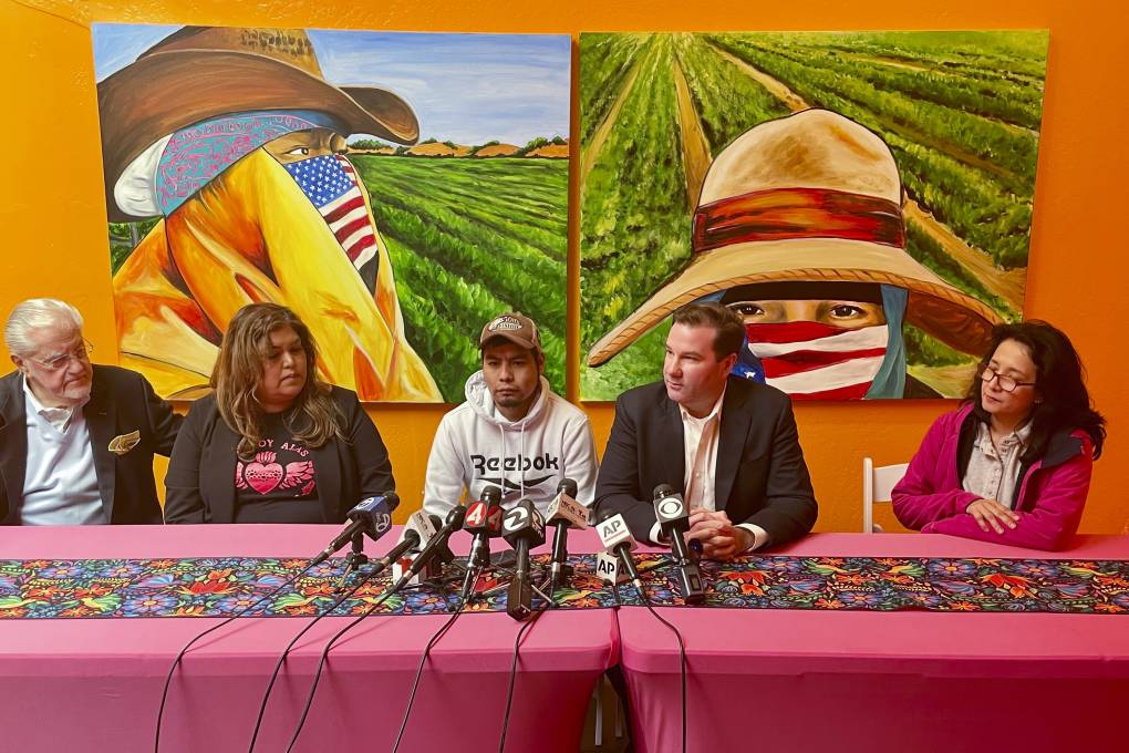 A young immigrant man of Latino origin sits between a woman and a man with microphones in front on a desk and a mural depicting immigrant farmworkers on the wall behind.