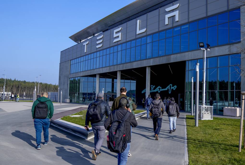 Several people wearing backpacks walk into a building that says "Tesla" on the front.