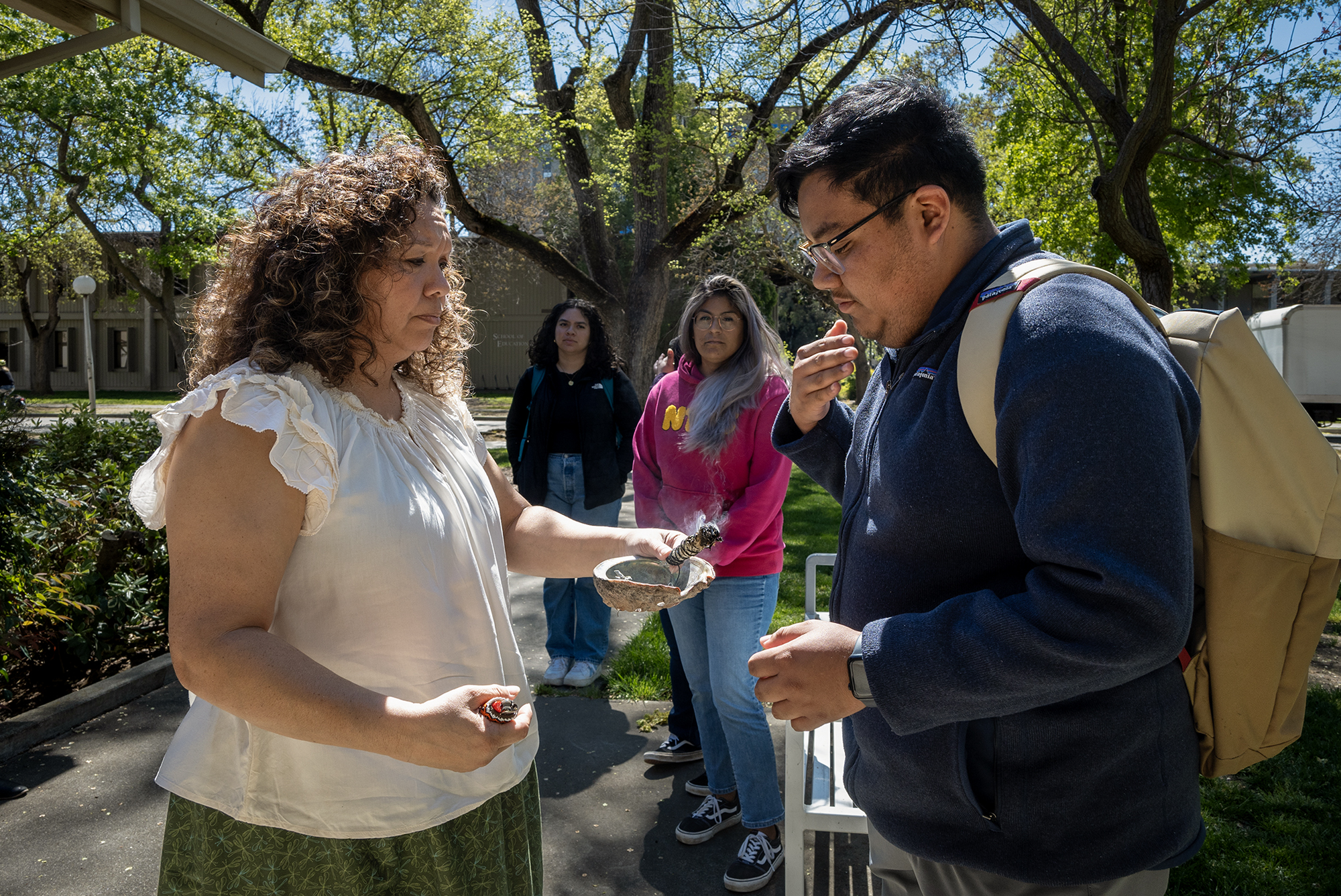 A native American woman in a white dress and a Native American man in a blue shirt and two people in the background during a ceremony.