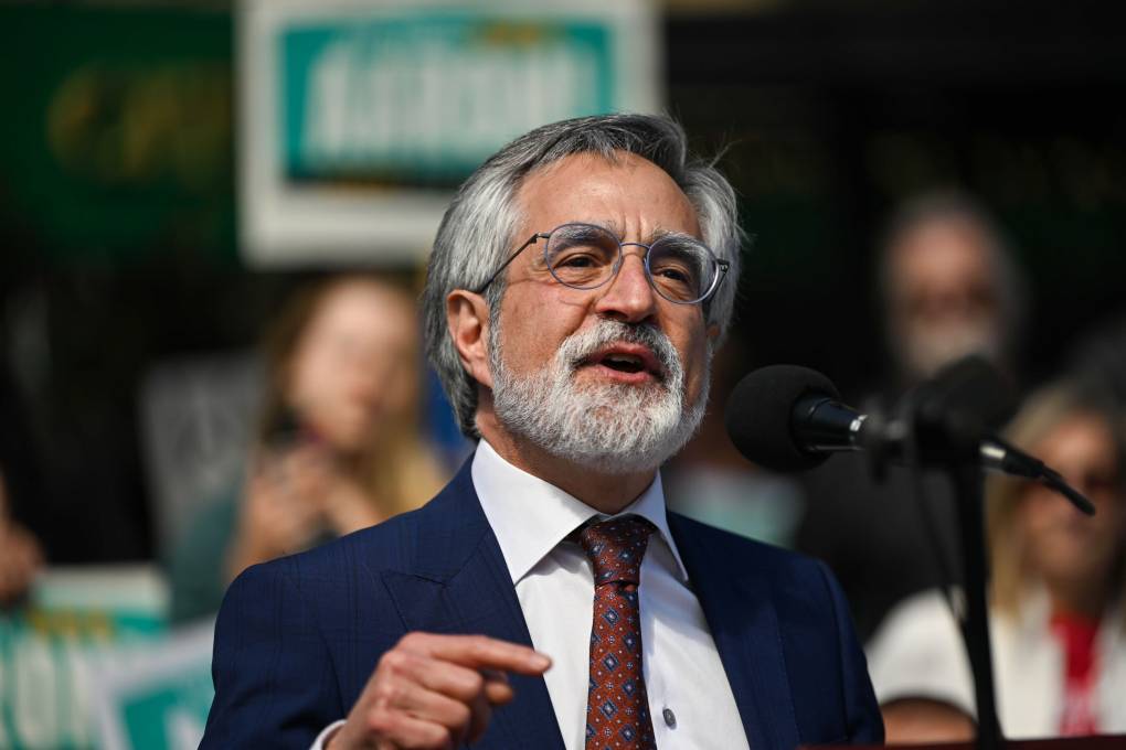 A middle aged man with whitened hair and beard and glasses wearing suit and tie speaks with blurred supporters behind him holding green signs.