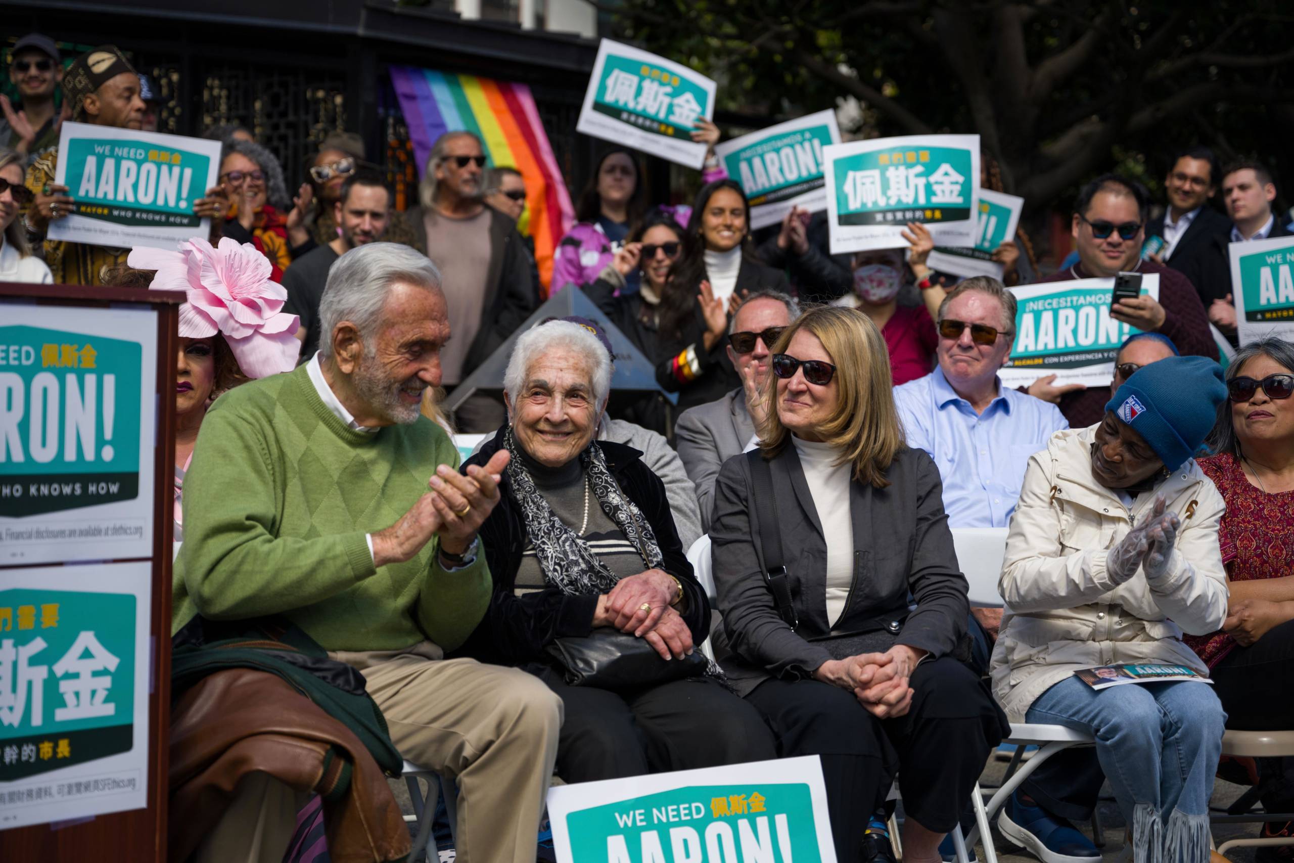 An older lady sits with people on either side and supporters of Aaron Peskin behind her waving signs.