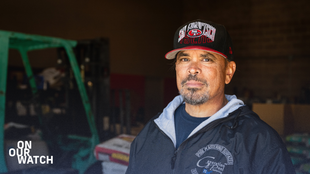 A Latino man, middle-aged with a graying goatee, gazes directly into the camera amidst a warehouse backdrop. He sports a black and red San Francisco 49ers cap and a dark blue windbreaker jacket adorned with a logo reading 'Generation Pool Plastering.' In the blurred background, a green warehouse vehicle and various boxes and supplies are visible.