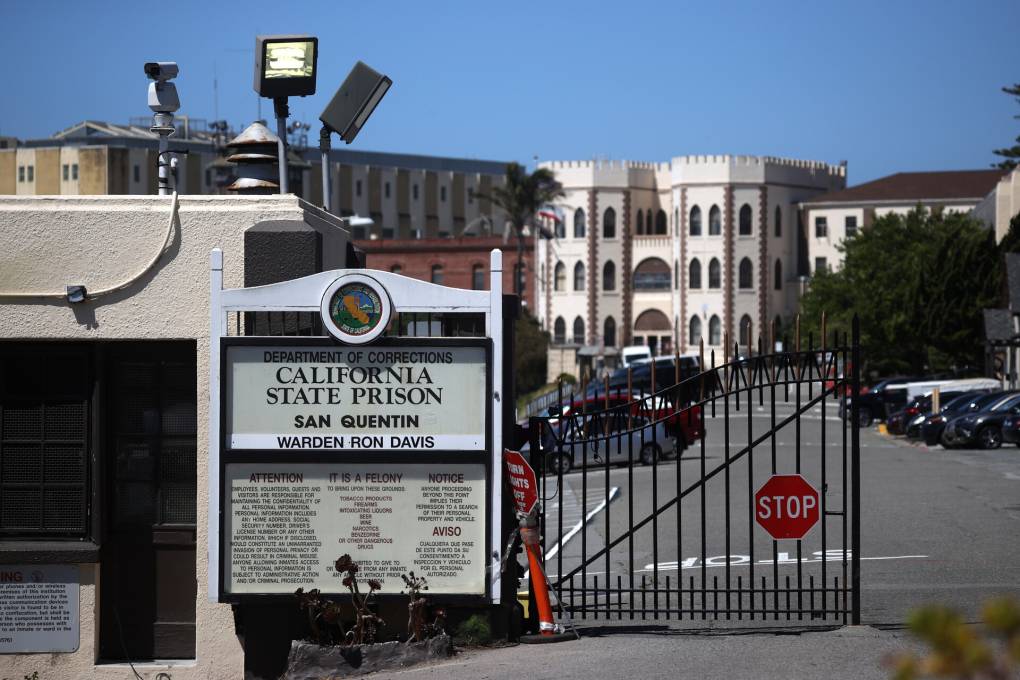 A view of a sign that reads 'California State Prison San Quentin' next to a gate with a prison in the background.