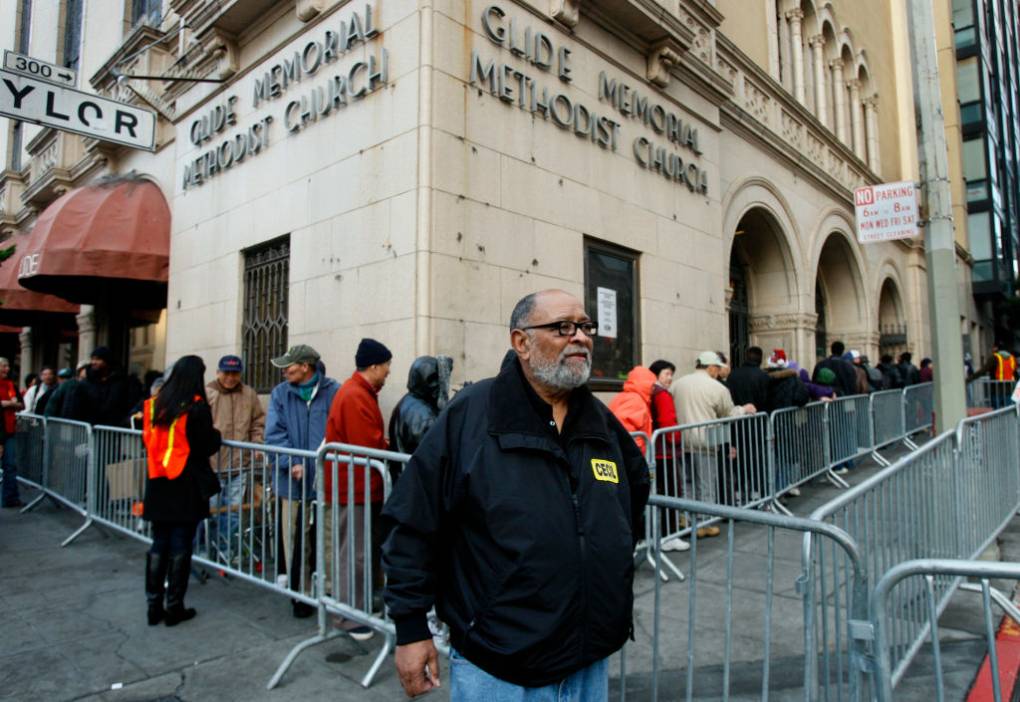 An older Black man walks outside Glide Memorial Methodist Church, where barricades are set up and people stand in line.
