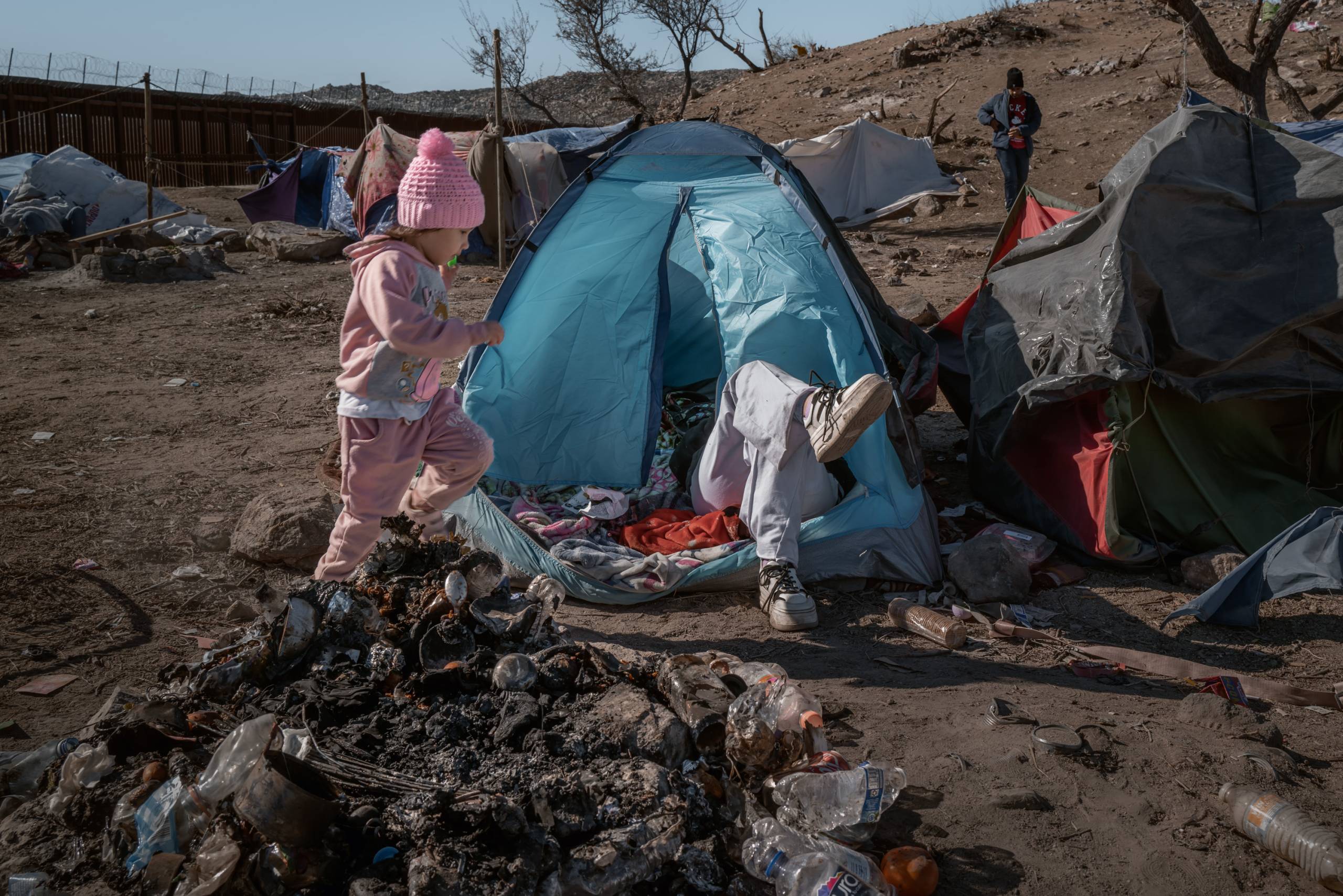 a little girl runs past trash in front of dilapidated tent in the desert, with more tents and trash in the background