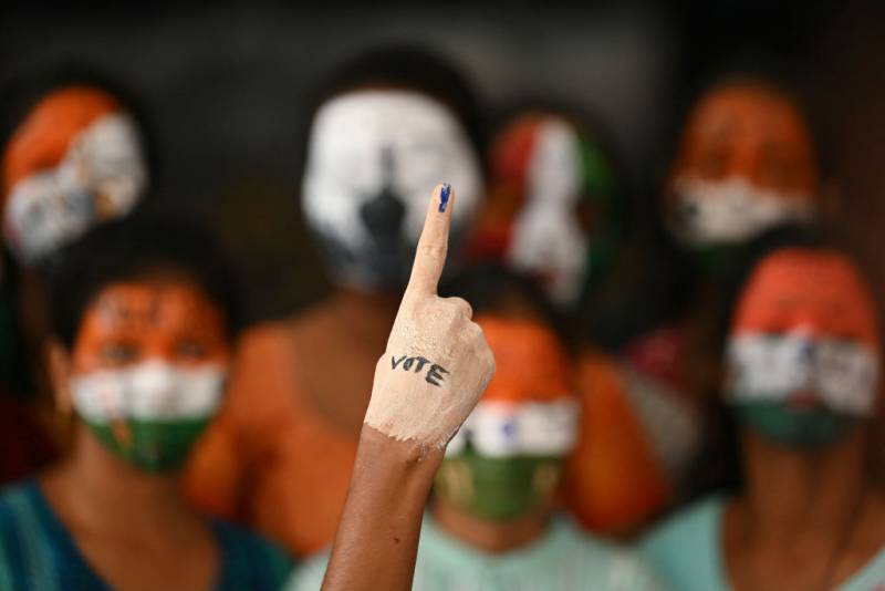 An arm with an index finger points upward. Ink is on the fingertip, and the word "vote" is written on the back of the hand. Blurred faces painted with Indian flags are seen in the background.
